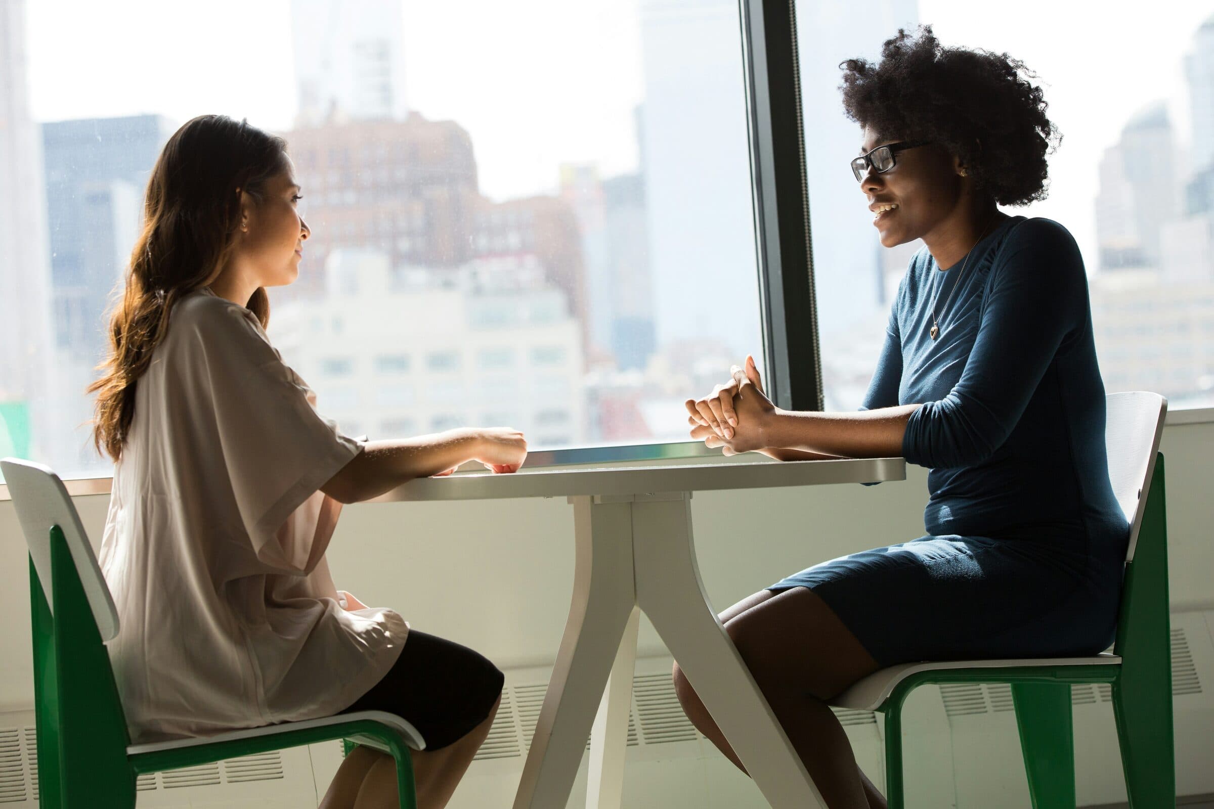 Two people in conversation across a table beside a bright window.